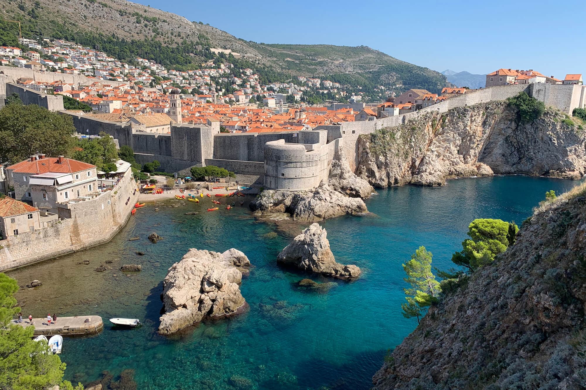 Medieval city wall of Dubrovnik, along with a small beach and kayakers enjoying the crystal-clear waters.