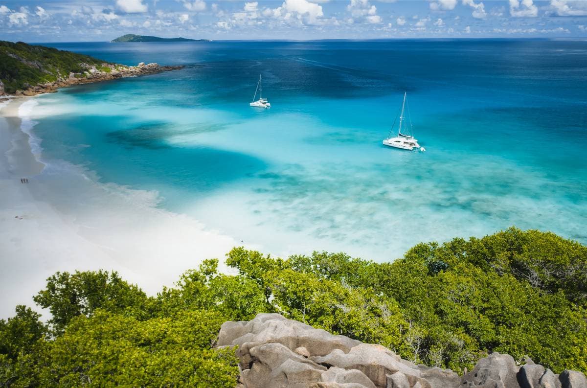 Aerial view of Grand Anse Beach on La Digue Island, Seychelles, featuring a stunning white sandy beach and a vibrant blue ocean lagoon, with catamaran yachts anchored.