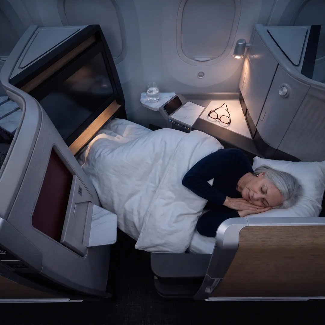 Seen from above: Linda asleep under a white duvet in her lie-flat Business Class bed, the cabin dark, her glasses folded beside a small reading light